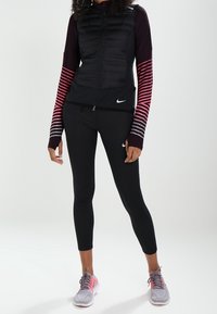 Sportovní outfit Nike zahrnující černou péřovou vestu, dlouhý pruhovaný top v purpurové barvě, černé legíny a šedobéžové sneakers.