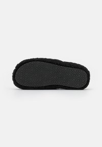 Semelle de pantoufle noire avec une prise texturée présentant des motifs en losange et circulaires, bords doublés d’un matériau doux et duveteux.