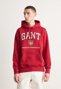 Sudadera roja de algodón, con un bolsillo frontal, capucha con cordón y texto impreso que incluye "GANT" y "AMERICAN SPORTSWEAR."