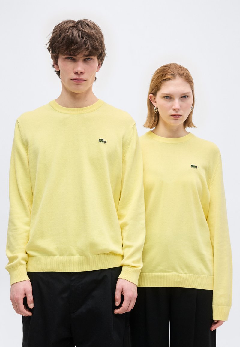 Νέος άνδρας και γυναίκα όρθιοι δίπλα-δίπλα, φορούν ταιριαστά ανοιχτό κίτρινα πουλόβερ Lacoste και μαύρο παντελόνι, με φόντο λευκό.