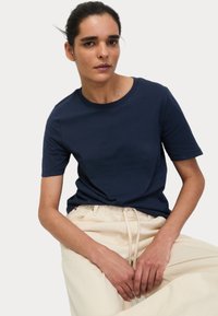 T-shirt in cotone blu navy con scollo rotondo e maniche corte, abbinata a pantaloni beige chiaro con cordoncino, che mostrano una texture liscia e un design semplice.