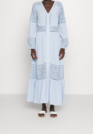 Žena v dlouhých světle modrých maxi šatech s krajkovými panely, zapínáním na knoflíky vpředu a bílými nazouvacími sandály.