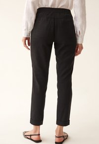 Zwarte, elastische taillebroek met een tapered fit en opgerolde boorden. Heeft achterzakken en een zachte, gestructureerde stof. Gedragen met platte sandalen.