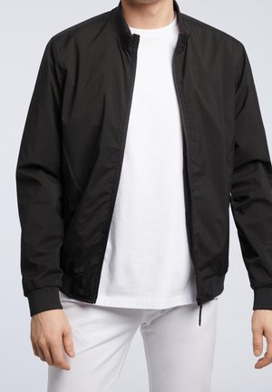 Mężczyzna ubrany w czarną kurtkę na suwak na białej koszulce i białych spodniach, stojący na tle jasnego, jednolitego tła.