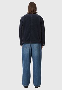 Personne aux cheveux bruns mi-longs portant une veste en velours noire et un jean bleu ample, debout de dos sur un fond gris clair.