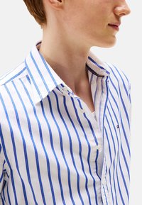 Camicia bianca con bottoni, caratterizzata da strisce verticali blu, un colletto classico e un piccolo logo sul petto. Tessuto di cotone morbido.