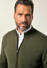 Hombre con cabello corto y oscuro y barba, vistiendo un suéter verde con cremallera sobre una camisa blanca de cuello, mirando directamente con expresión seria.