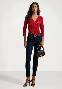 Egy nő piros mintás felsőt visel, sötét skinny farmert, fekete övet, fekete magassarkú szandált, és egy fekete kézitáskát tart, arany kapoccsal.