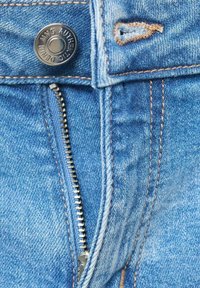 Podrobnosti denim hlač, ki vključujejo srebrno zadrgo, strojno obarvano rjavo šivanje in blagovno znamko kovinskega gumbka. Svetlo modra tkanina z rahlo obrabo.