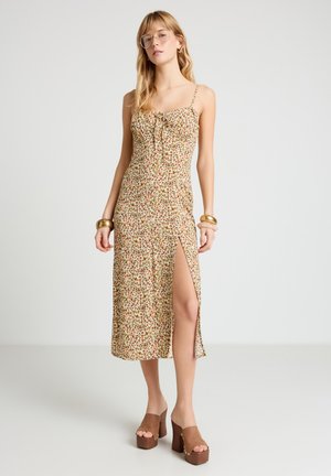 Donna che indossa un vestito midi floreale con spalline sottili, spacco laterale, grandi bracciali dorati e sandali con zeppa marroni, in piedi su uno sfondo chiaro.