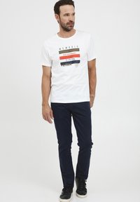 T-shirt blanc avec des graphiques de texte multicolores et un col rond, associé à un pantalon slim-fit bleu marine et des baskets noires.