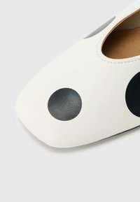 Balti odiniai batai su smailia nosimi, didelėmis juodomis polka taškeliais ir atidengtu šonu. Lygi tekstūra su minimaliais siūlais.