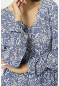 Blouse bleu à motif floral avec manches longues froncées et encolure en V. Le tissu a une finition texturée, avec des motifs de feuilles blanches.