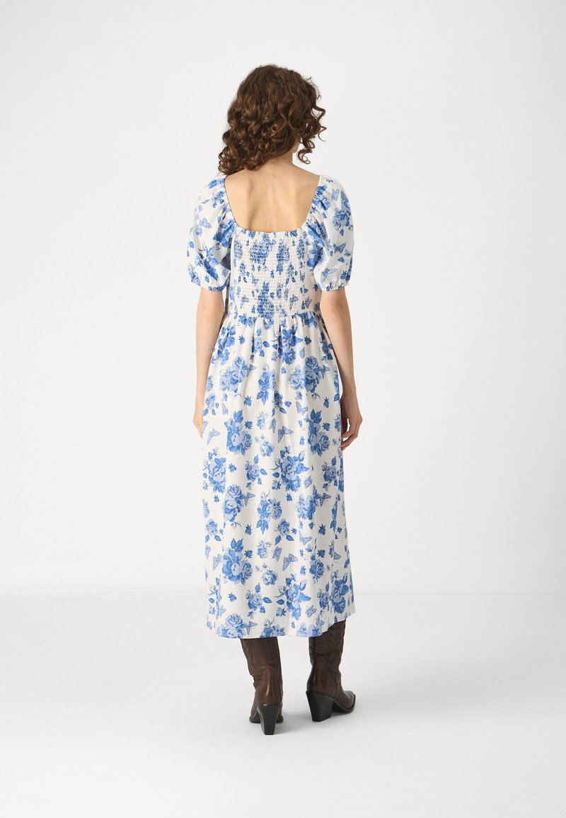 Robe maxi florale blanche avec des motifs bleus. Elle présente des manches bouffantes et un dos carré. Assortie à des bottes à talons brunes. Texture de tissu lisse.