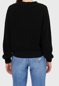 Personne portant un pull en tricot noir et un jean en denim bleu clair, vu de dos sur un fond clair et uni.