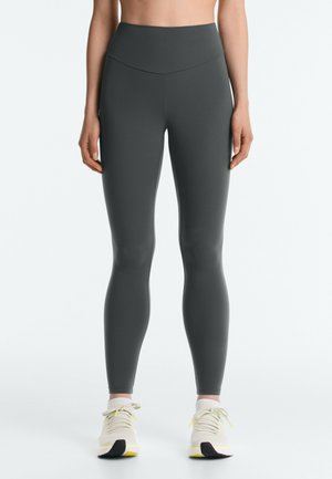 Kvinde iført mørkegrå højtaljede leggings og hvide træningssko, stående med armene afslappet langs siden på en ensfarvet baggrund.