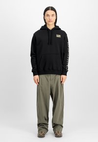 Sudadera negra con capucha y un bolsillo tipo canguro en la parte delantera, que presenta un parche de camuflaje y texto blanco en la manga. Combinada con pantalones de cargo holgados en color oliva.