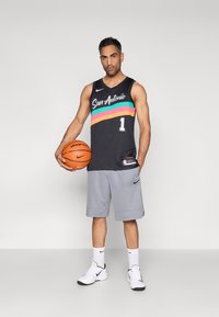 Tricou negru de baschet cu inscripția „San Antonio” și dungi colorate, asortat cu pantaloni scurți gri, ținând un coș portocaliu, purtând adidași albi și șosete albe.