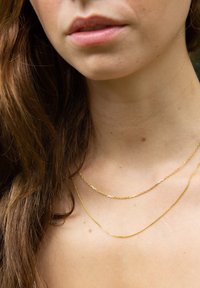 Goldfarbene Halsketten mit einem dünnen, gedrehten Design, die um den Hals geschichtet sind. Die Haut zeigt eine glatte Textur, mit sichtbaren Sommersprossen in der Nähe des Schlüsselbeins.