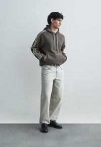 Καφέ φούτερ με κουκούλα και λευκές ρίγες στη πλευρά, συνδυασμένο με ανοικτόχρωμο τζιν και μαύρα παπούτσια. Απλός σχεδιασμός με άνετη εφαρμογή.