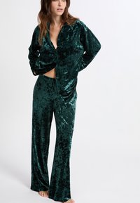 Sada sametových pyžam v emeraldově zelené barvě s košilí s dlouhým rukávem na knoflíky a širokými kalhotami s jemnou, lesklou texturou.