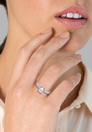 Diamantring mit rechteckigem Mittelstein, silbernem Ringband und kleineren Diamanten entlang des Ringbands, getragen an einer Hand in der Nähe des Gesichts.