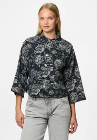 Vrouw in een zwarte cropped jas met grijze bloemenpatronen en driekwartmouwen, gecombineerd met lichtgrijze high-waisted jeans.