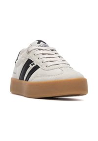 Zapatilla de ante gris con rayas negras, punta redondeada y suela de goma. Presenta cordones y logotipo en la lengüeta. Diseño plano, estilo minimalista.