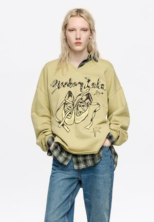 Sudadera oversized de color verde oliva con escote en V y un gráfico de zapatos. Debajo, se asoma una camisa de cuadros verde oscuro. Jeans de color azul claro.