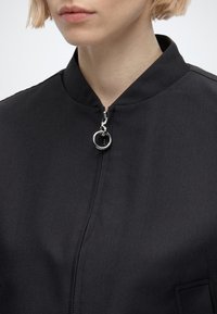 Gros plan d’une personne portant une veste noire avec un tirette de fermeture éclair en anneau argenté et un col court montant.