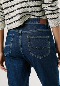 Jeans in denim blu scuro con vita alta, dotati di tasche posteriori con dettagli cuciti e un'etichetta in pelle marrone sulla vita.