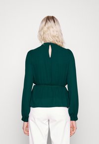 Blusa verde a maniche lunghe con dettaglio a chiave sul retro, vita cintrata e tessuto fluido, abbinata a pantaloni bianchi.