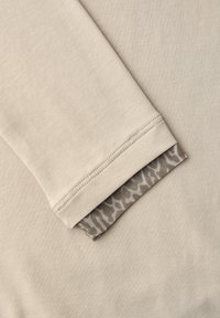 Beige long-sleeve shirt featuring a textured cuff with a geometric-patterned mesh lining, highlighting contrasting materials and simple design.