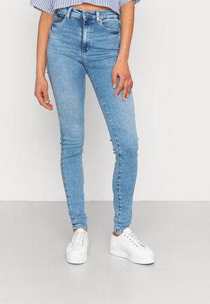Vrouw draagt lichtblauwe skinny jeans en witte plateau sneakers, staand tegen een effen lichtgrijze achtergrond.