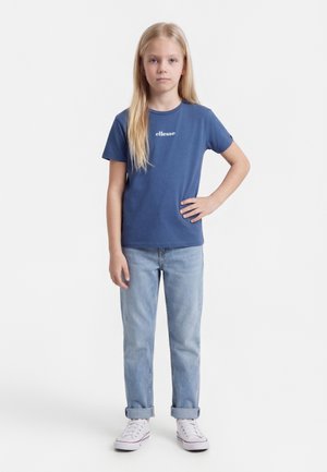 Niña con cabello largo y rubio, de pie mirando hacia adelante, vistiendo una camiseta azul de ellesse, jeans azul claro con puños y zapatillas blancas.