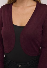 Bordeaux gebreide cardigan met een v-halsontwerp, lange mouwen en een glinsterende textuur. Draag het over een zwarte top, wat een aangesloten silhouette laat zien.