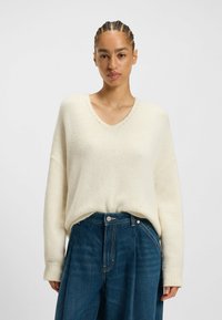 Kremowy sweter z dekoltem w szpic, z ryflowanym wykończeniem rękawów i subtelnymi detalami szycia, zestawiony z wysokim stanem niebieskimi szerokimi spodniami dżinsowymi.