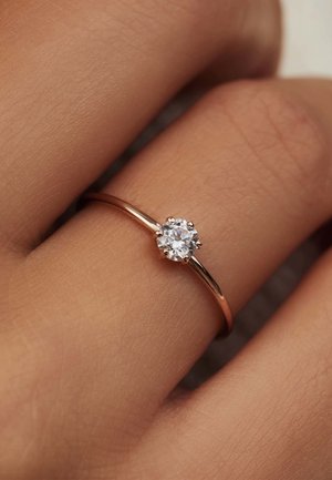 Rose gold ring with a round, clear stone set in six prongs; smooth, polished band; worn on a hand with a textured skin surface.