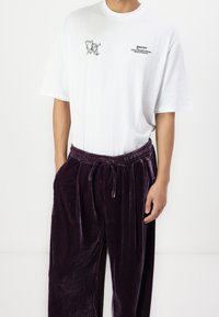 Vit bomull t-shirt med ett grafiskt tryck på vänster bröst, ihop med lila sammetbyxor som har resårmidja och dragsko.