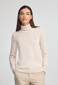 Maglione beige a collo alto realizzato in morbido materiale a maglia, con un colletto e polsini a coste, e un piccolo dettaglio del logo sul petto.