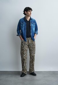 Sötét, göndör hajú férfi, kék farmeringben, fekete V-nyakú pólóban, leopárdmintás nadrágban és fekete cipőben, zsebre tett kézzel áll.