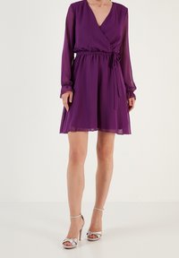 Žena v dlouhých rukávech fialové zavinovací šaty s uvázaným pasem, v kombinaci se stříbrnými sandály na vysokém podpatku s páskem kolem kotníku, stojící na bílé podlaze.