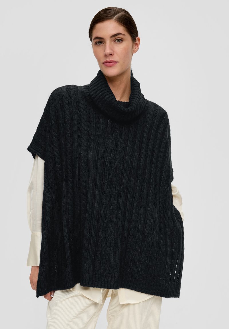 Zwarte gebreide poncho met een hoge geribbelde kraag, voorzien van een kabelbreipatroon en korte mouwen, gedragen over een lichtgekleurde shirt.