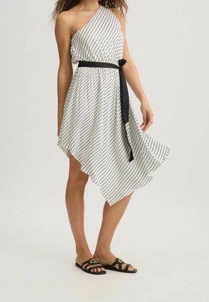 Kobieta w białej sukience na jedno ramię z czarnymi ukośnymi paskami, asymetrycznym dołem, czarnym paskiem w talii i czarnych sandałach na odkrytej stopie.