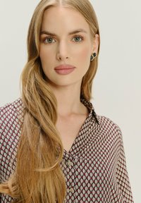 Brązowa i kremowa bluzka w wzór z kołnierzykiem, z geometrycznym designem i guzikami z przodu. Kobieta z długimi blond włosami.