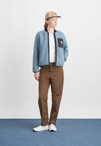 Kék fleece dzseki fekete zsebbel, barna nadrág és fehér sneaker. A dzsekin cipzár található, és laza szabású.