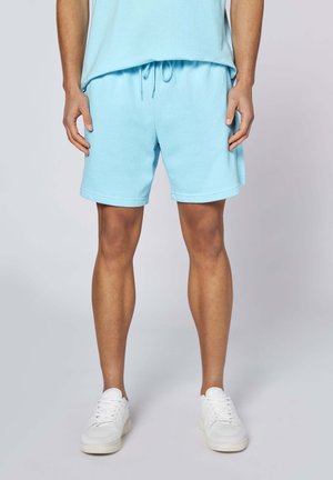Mann trägt hellblaue Shorts mit Kordelzug und passendem Hemd, steht mit weißen Sneakers vor einfachem Hintergrund.
