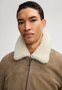 Giacca in suede marrone con colletto in shearling color crema, dotata di zip frontale e texture liscia. Primo piano sul volto del modello.