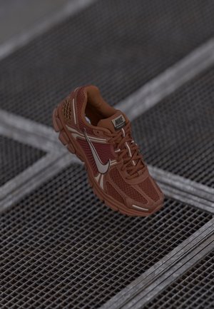 Bruine Nike Vomero 5 hardloopschoen met een bovenwerk van mesh en synthetisch materiaal, gedetailleerde witte swoosh, zittend op een metalen roostervloer.