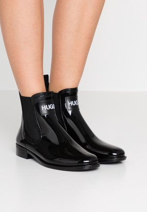 Błyszczące czarne botki za kostkę z elastycznymi panelami po bokach, niskimi obcasami i logo "HUGO" z przodu, noszone na gołych nogach na jednolitym tle.